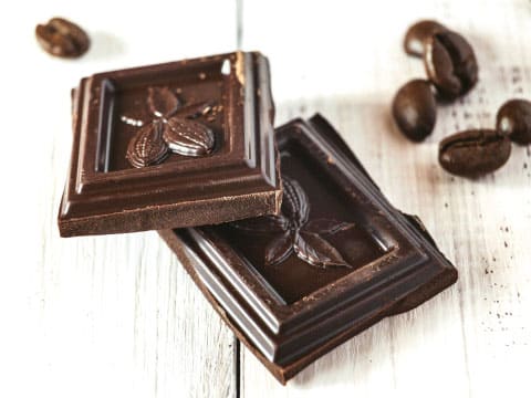 Two dark chocolate squares with a leaf-embossed design, resting on a white wooden surface surrounded by scattered whole coffee beans