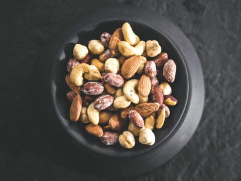 An overhead view of a black bowl filled with salted mixed nuts — almonds, cashews, hazelnuts, and pistachios — on a dark stone surface