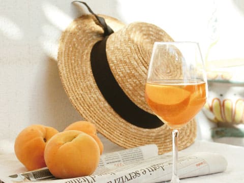 A glass of orange-hued apricot wine beside a straw hat and fresh whole apricots, evoking a relaxed summer mood