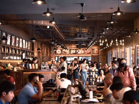 Customers dining and ordering at a bustling cafe with wooden interiors, pendant lighting, and a service counter