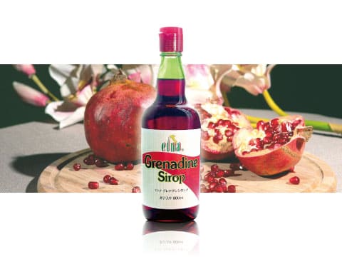 A bottle of Etna grenadine syrup standing on a wooden board surrounded by fresh pomegranates and flowers