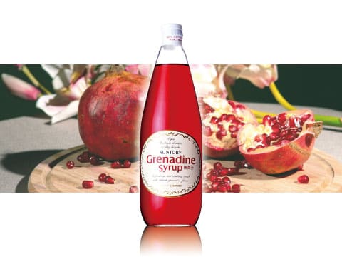 A bottle of Suntory grenadine syrup standing on a wooden board surrounded by fresh pomegranates and flowers