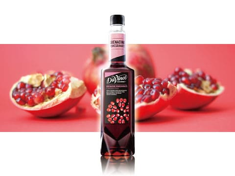 A bottle of DaVinci grenadine pomegranate syrup standing between halved pomegranates against a red background