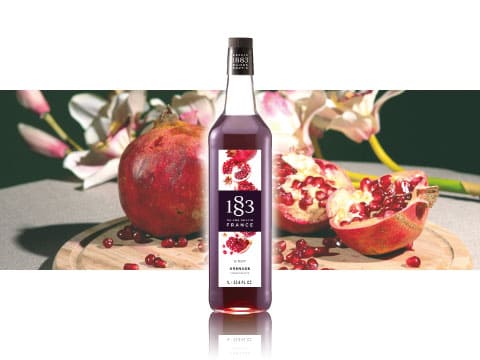 A bottle of Maison Routin 1883 grenadine syrup standing on a wooden board surrounded by fresh pomegranates and flowers