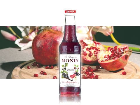 A bottle of Monin grenadine syrup on a wooden board surrounded by fresh pomegranates, berries, and flowers