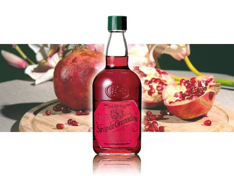 A bottle of Meidi-Ya grenadine syrup standing on a wooden board surrounded by fresh pomegranates and flowers