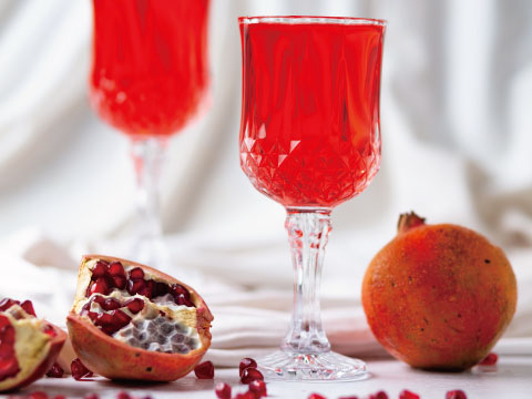 Two crystal glasses filled with a bright red pomegranate drink, beside a halved pomegranate with visible seeds and a whole pomegranate