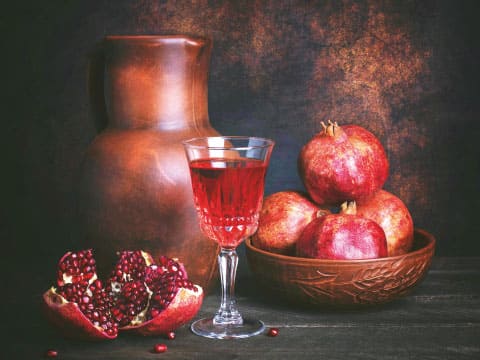 A still life of fresh pomegranates, one cut open, beside a glass of red liquid and a ceramic pitcher