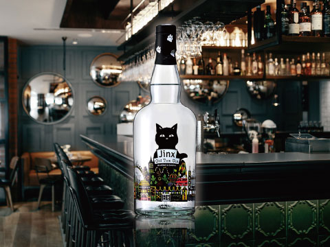 Bottle of Jinx Old Tom Gin with a black cat and colorful cityscape label standing on a bar counter, with round mirrors and bar stools behind