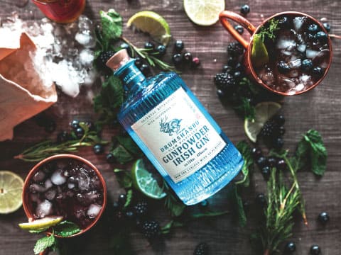 A bottle of Drumshanbo Gunpowder Irish Gin surrounded by two copper mugs of blackberry cocktails, fresh herbs, lime wedges, and crushed ice on a wooden table