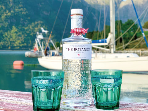 A bottle of The Botanist Islay Dry Gin with two green glasses on a wooden table, set against a scenic harbor with sailboats