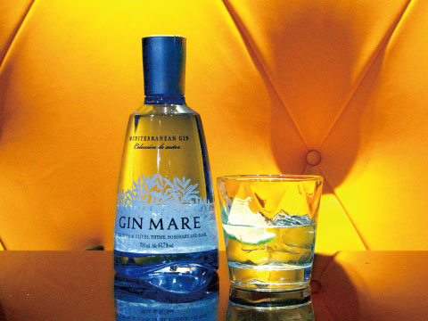A bottle of Gin Mare Mediterranean Gin beside a glass of gin and tonic with lime, set against a vivid yellow tufted background