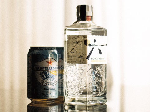 Bottle of Roku Japanese Craft Gin with engraved cherry blossom patterns standing beside two cans of tonic water on a reflective table