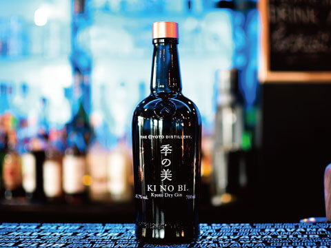 Dark bottle of Ki No Bi Kyoto Dry Gin with a copper-colored cap standing on a bar counter with softly blurred blue lights behind