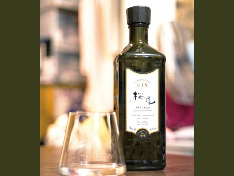 Dark green bottle of Sakurao Japanese Dry Gin with a cream label standing beside an empty tasting glass on a wooden table
