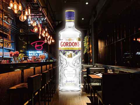 A large bottle of Gordon's London Dry Gin displayed prominently in a moody bar interior with warm string lights