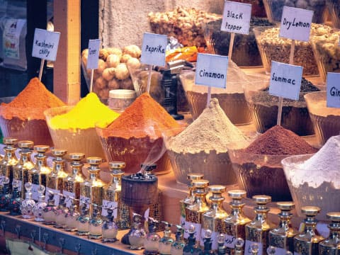 Market stall with bowls of colorful ground spices labeled Kashmiri chili, paprika, cinnamon, black pepper, dry lemon and sumac, above rows of glass bottles