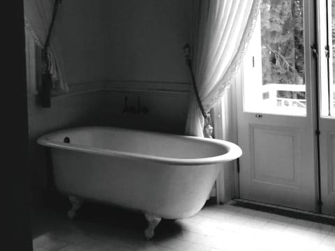 Black and white photo of a vintage clawfoot bathtub beside a window with sheer curtains