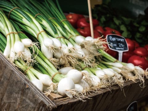 Bunches of fresh white pearl onions with green tops and roots displayed in a wooden market crate, with tomatoes in the background