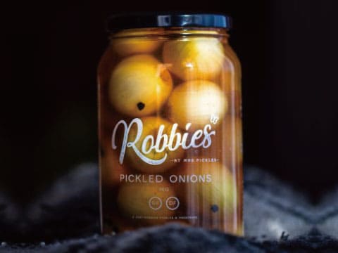A glass jar of pickled pearl onions labeled "Robbies Pickled Onions," set against a dark background