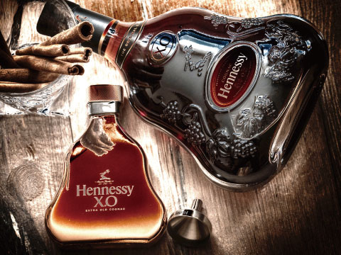A bottle of Hennessy X.O cognac beside its ornate crystal decanter and cinnamon sticks, displayed on a rustic wooden table