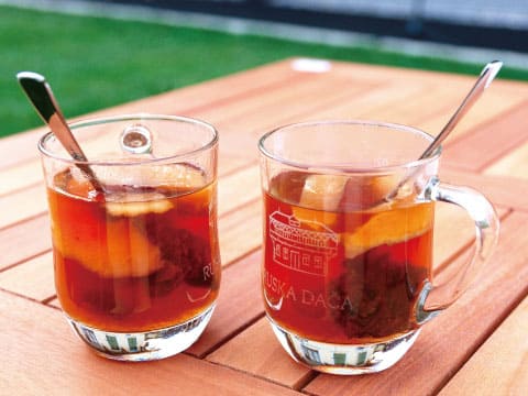 Two glass mugs of hot amber Grog cocktail with citrus slices and spoons, served on a wooden outdoor table with green grass in the background