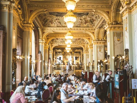 Guests dining in a grand, ornately decorated hotel hall with gold detailing, frescoed ceilings, and crystal chandeliers