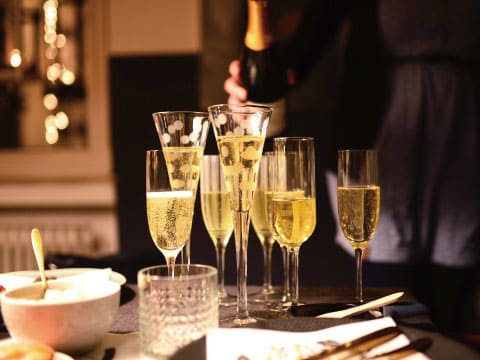 Champagne being poured into several flute glasses arranged on a table at an evening gathering, with warm string lights and a blurred figure pouring in the background