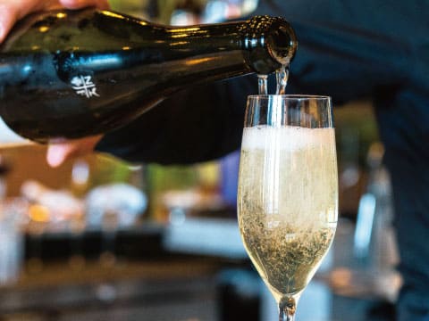 A bartender pouring sparkling wine from a dark bottle into a champagne flute, with fine bubbles rising and a softly blurred bar in the background
