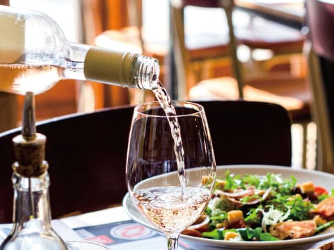 White wine being poured into a glass at a restaurant table, with a fresh salad and olive oil bottle nearby