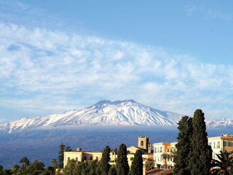 Snow-capped Mount Etna volcano rising above the Sicilian town of Taormina, with cypress trees and terracotta rooftops in the foreground