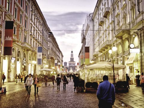 A bustling pedestrian street in Milan, Italy, lined with historic buildings and shops, with the Sforza Castle visible in the distance at dusk