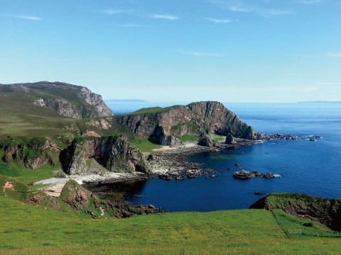A rugged coastline on the Isle of Islay, Scotland, with grassy cliffs overlooking a sheltered blue bay under a clear sky