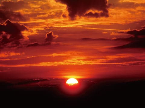 A dramatic red and orange sunset with the sun dipping below the horizon, silhouetted clouds and hills framing the sky