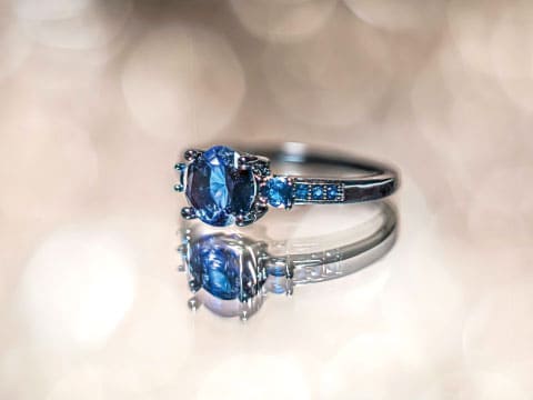 A blue sapphire ring surrounded by smaller sapphire accents, reflected on a mirrored surface with a softly blurred bokeh background