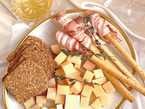 A platter with prosciutto-wrapped breadsticks, cubed cheese, and slices of seeded bread, garnished with fresh thyme