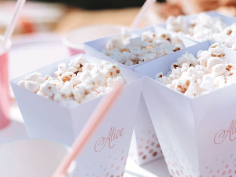 Buttered popcorn served in white paper cups with pink polka-dot accents and a pink striped straw, on a party table