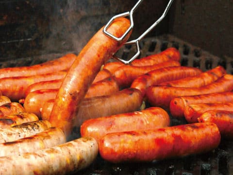 Assorted sausages grilling on a barbecue, with tongs lifting one sausage from the grate