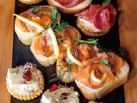 An assortment of smoked salmon canapés with lemon curls and dill, plus cream cheese and pomegranate crackers, arranged on a dark slate board