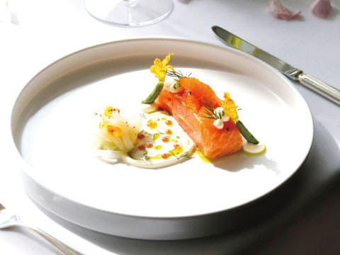 Fillet of salmon on a white plate, garnished with yellow edible flowers and dill, served with cream sauce, shaved vegetables and orange sauce dots