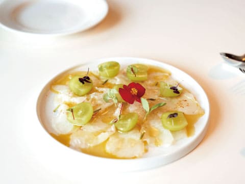 Thin slices of white fish carpaccio drizzled with citrus dressing, garnished with green tomatoes, an edible red flower, and herbs on a white plate