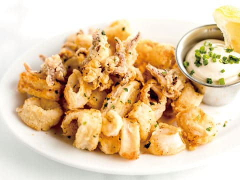 A white plate of golden fried calamari rings and tentacles, served with a small dish of creamy aioli garnished with chives and a lemon wedge