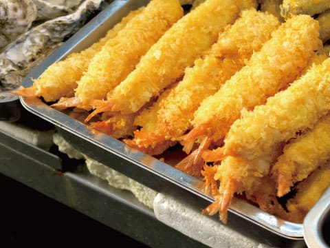 A metal tray lined with golden breaded fried shrimp, tails exposed, served on crushed ice alongside oysters