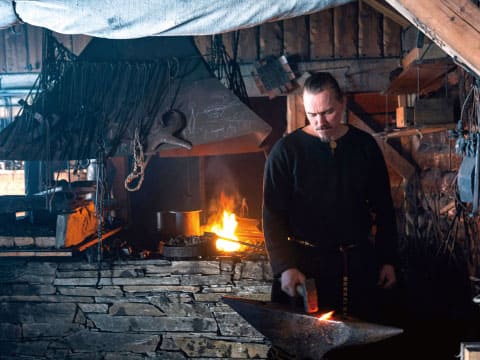 Blacksmith in a black tunic hammering glowing metal on an anvil next to a burning forge in a rustic workshop