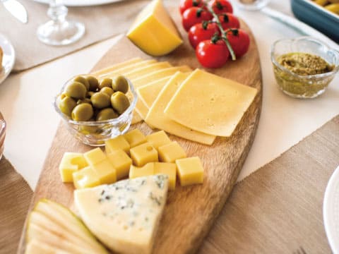 A wooden cheese board with sliced and cubed cheese, blue cheese, green olives, cherry tomatoes, and a small dish of mustard