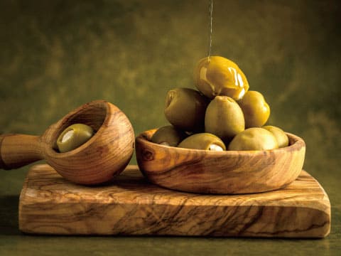 Green olives in a wooden bowl and spoon, with olive oil being drizzled over them on a rustic wooden board
