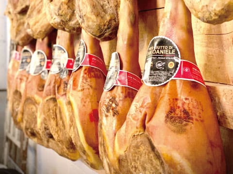 Rows of whole cured ham legs, labeled "Prosciutto di San Daniele," hanging in a curing room