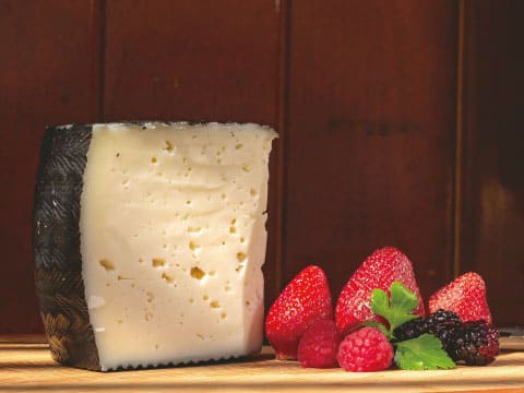 A wedge of Manchego cheese with its distinctive rind, served alongside fresh strawberries, raspberries, and blackberries on a wooden board