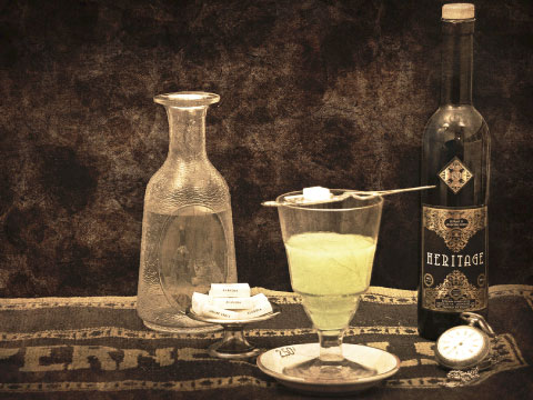 A glass of pale green louched absinthe with a sugar cube resting on an absinthe spoon, beside a water carafe and a bottle of Heritage absinthe on an antique table