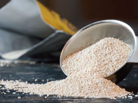 Granules of dry yeast spilling from a metal scoop onto a dark wooden surface beside an open package